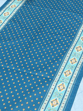 Load image into Gallery viewer, Acrylic coating allows for universal wipe clean finish, allowing for liquids and oils to bead off. Measures 16.5 by 60 in. Pictured on a 72 inch length table for reference, but may be draped over the sides of a smaller table as well. Manufactured by l'Ensoillade and made in France. Pictured in deep jade blue with light turquoise and golden yellow accents.