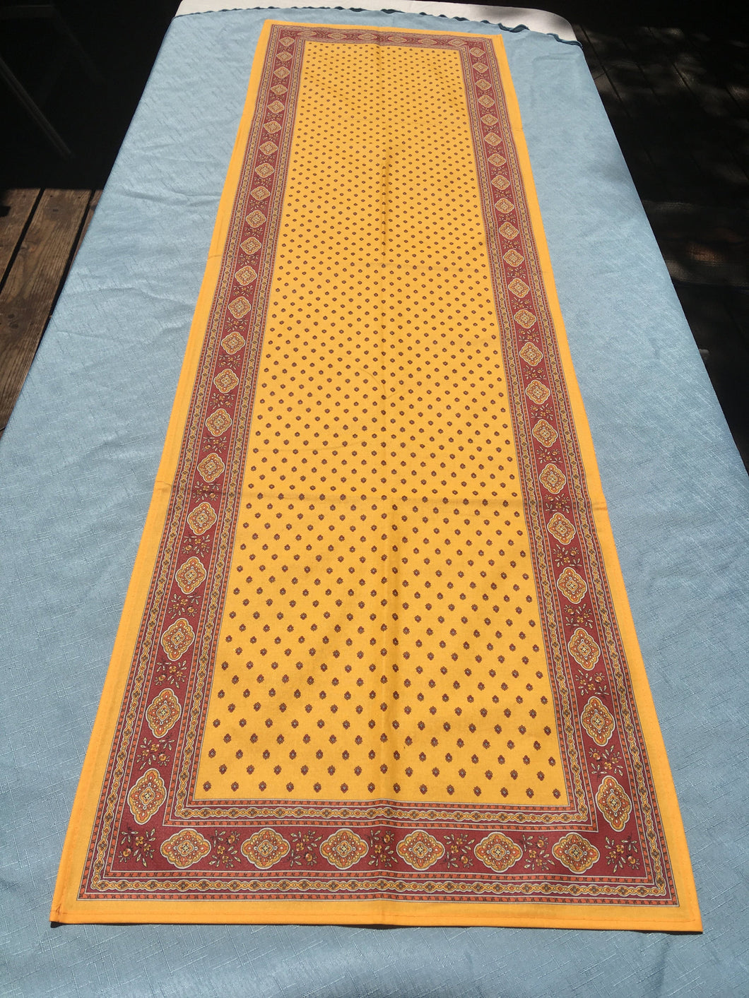 Acrylic coating allows for universal wipe clean finish, allowing for liquids and oils to bead off. Measures 16.5 by 60 in. Pictured on a 72 inch length table for reference, but may be draped over the sides of a smaller table as well. Manufactured by l'Ensoillade and made in France. Pictured in safran yellow with deep red accents.